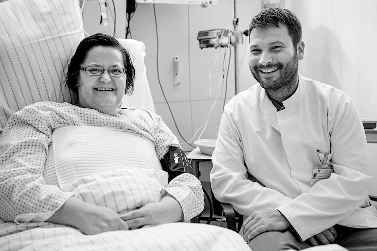 Foto eines Arztes, der am Krankenhaus-Bett einer Patientin sitzt, beide lächeln.