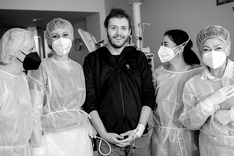 Foto eines Arztes, der am Krankenhaus-Bett einer Patientin sitzt, beide lächeln.